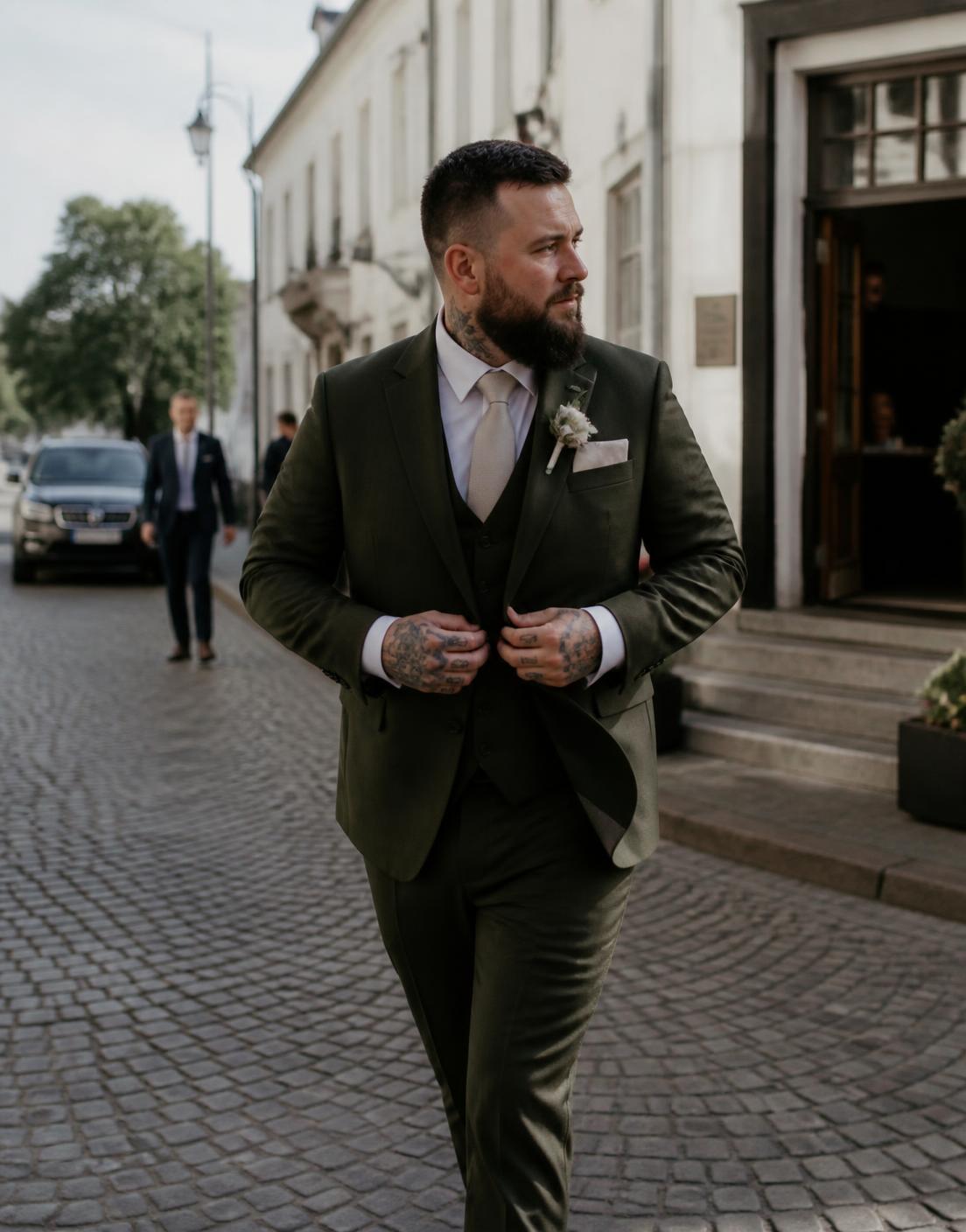 Bruidegom met baard en tattoos in een olijfgroen driedelig pak, lopend over een geplaveide straat terwijl hij zijn colbert dichtknoopt